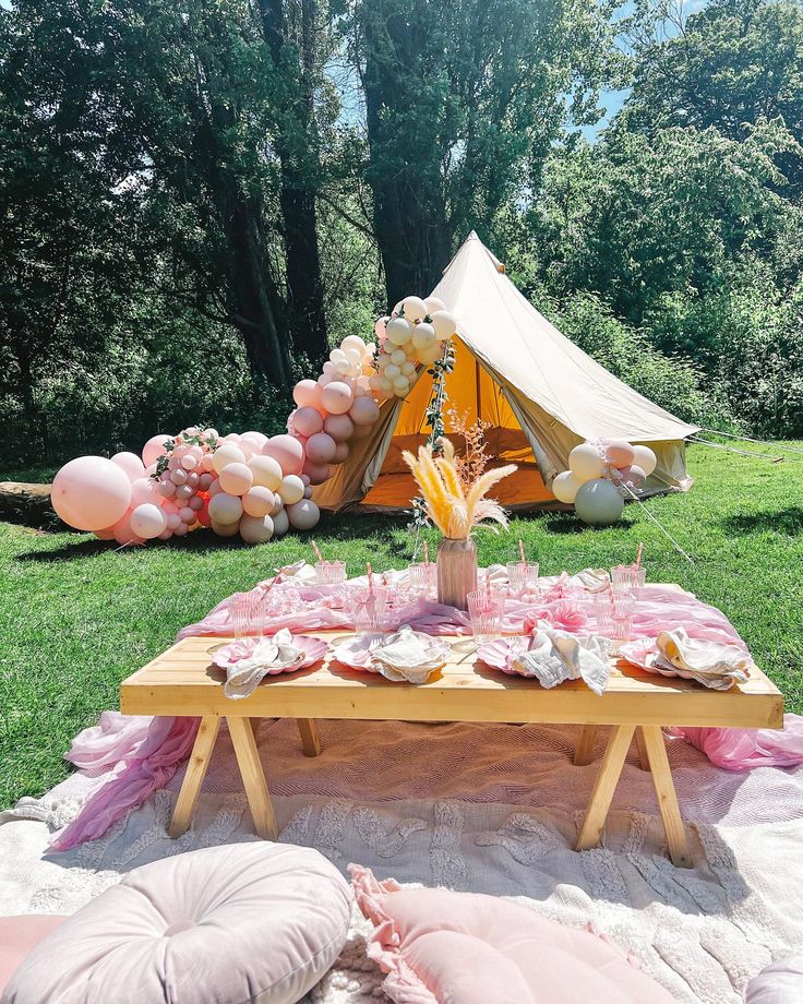 a table set up for a party with pink and white balloons on the grass, near a teepee tent