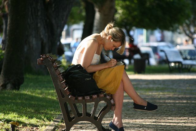 a woman is sitting on a park bench and writing in her notebook while looking down at her purse