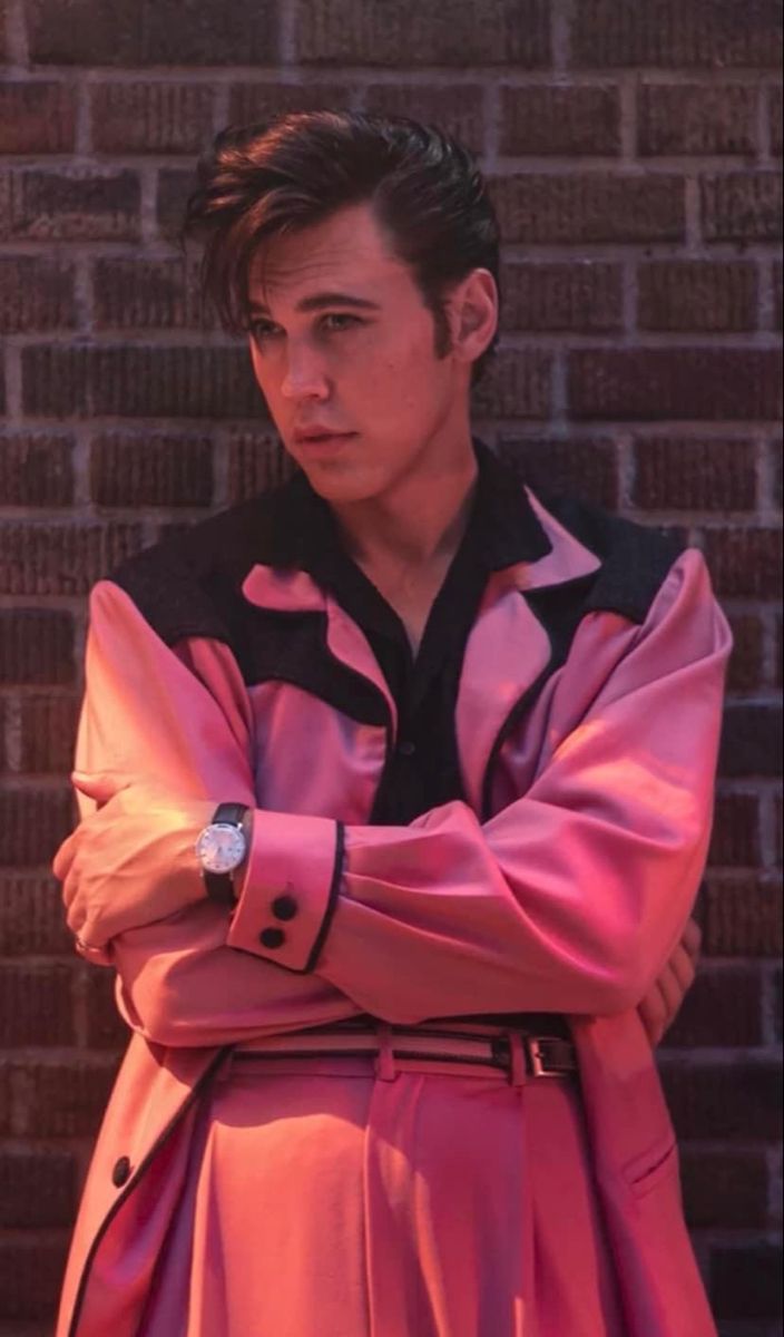 a man with his arms crossed standing in front of a brick wall wearing a pink trench coat