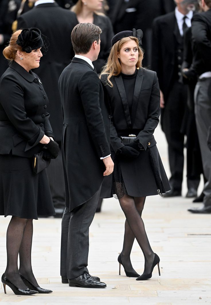 two women and a man in black are standing near each other on the street while people stand behind them