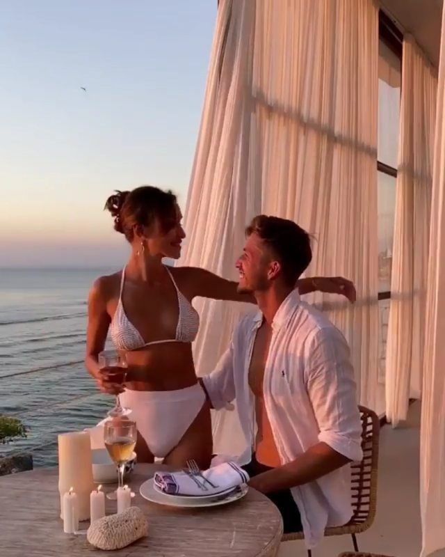 a man standing next to a woman at a table near the ocean with food and drinks