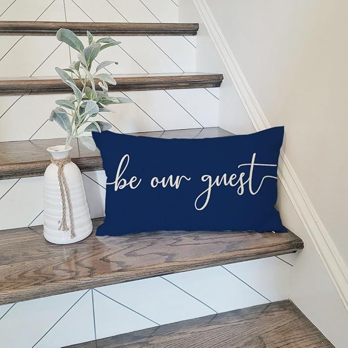 a blue pillow with the words be our guest on it next to a white vase