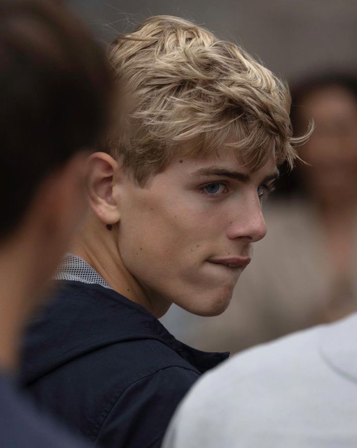 a young man with blonde hair and blue eyes looks to his left as he stands in front of other people