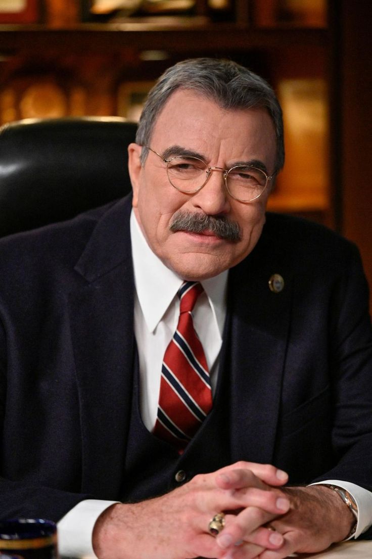 an older man in a suit and tie sitting at a desk with his hands folded