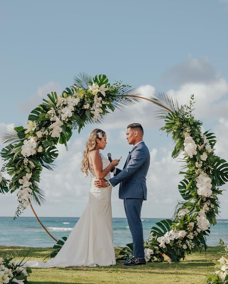 a bride and groom standing under an arch with white flowers on the grass by the ocean