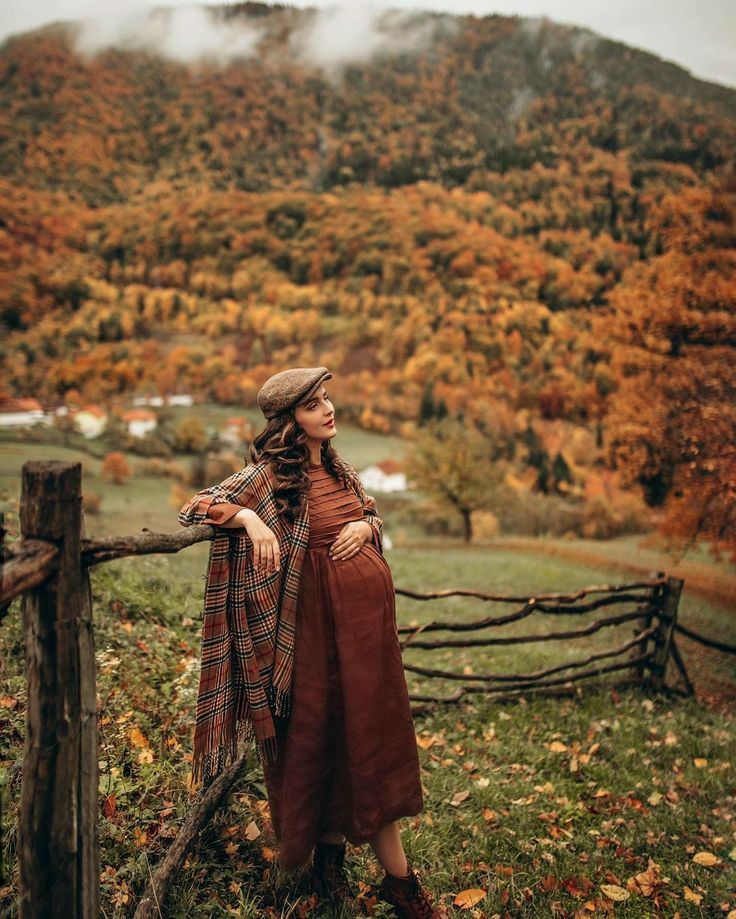 a woman standing on top of a lush green field next to a wooden fence covered in fall leaves