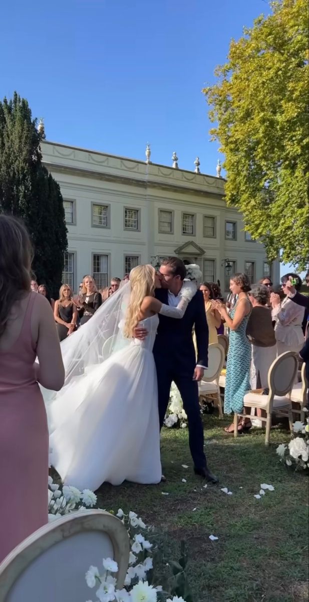 a bride and groom kissing in front of an outdoor wedding ceremony with white flowers on the grass