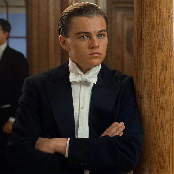 a young man in a tuxedo leaning against a wooden wall with his arms crossed