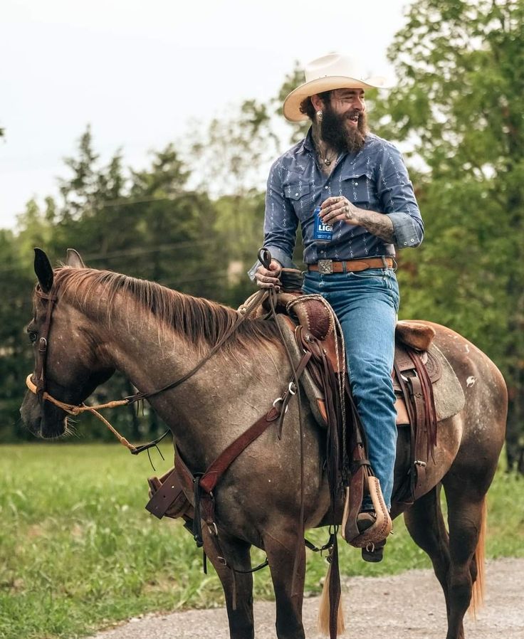 a man wearing a cowboy hat and jeans riding a brown horse on a dirt road