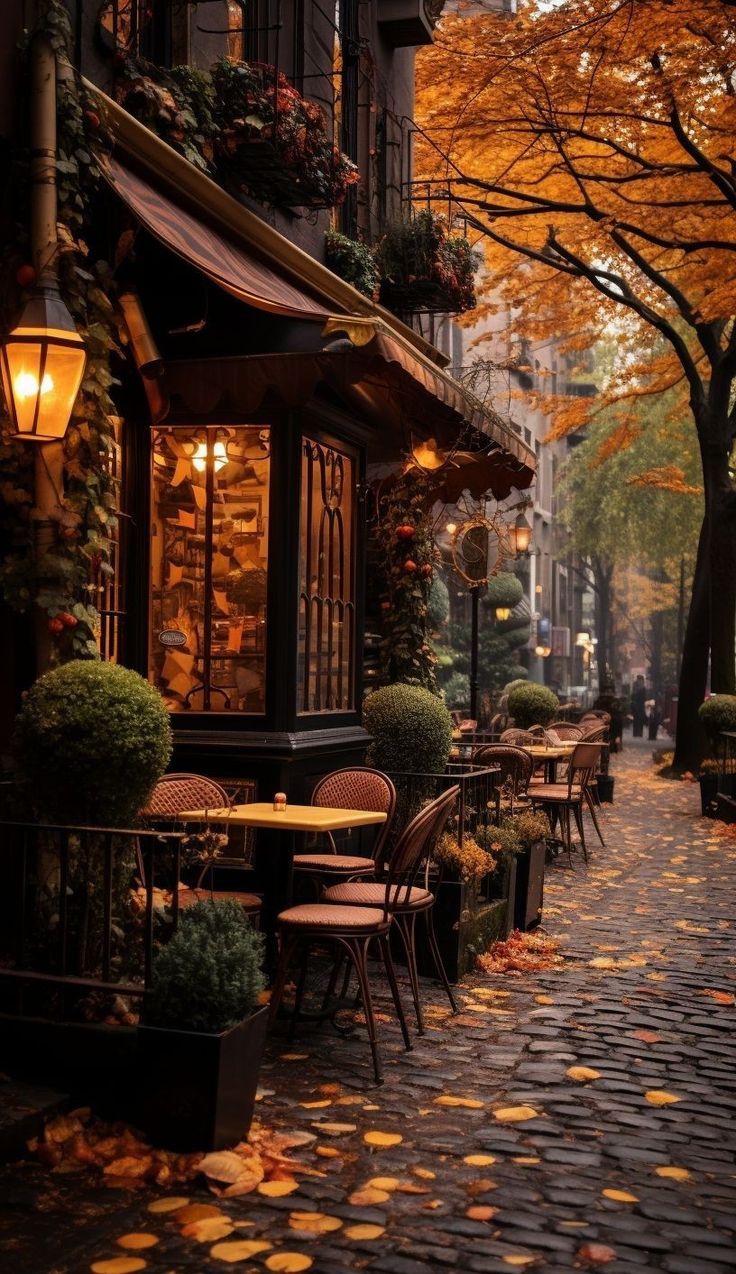 an empty street with tables and chairs on the sidewalk in front of trees that have fallen leaves