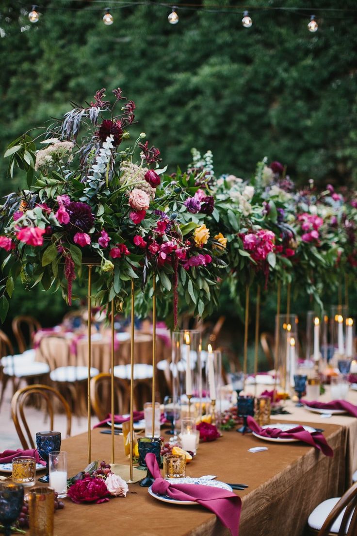 a long table topped with lots of purple and red flowers next to tall gold candles