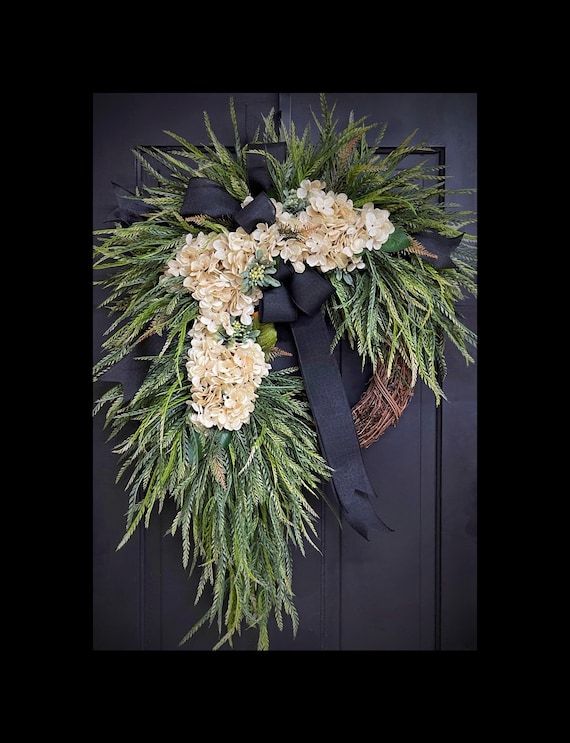 a wreath with white flowers and black ribbon hanging on a front door, decorated with greenery