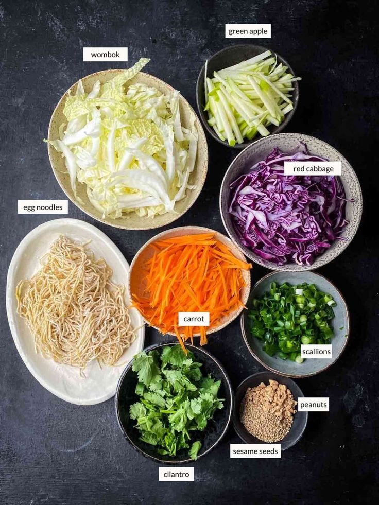 the ingredients for cabbage salad laid out in bowls on a black surface, including carrots, celery, lettuce and other vegetables