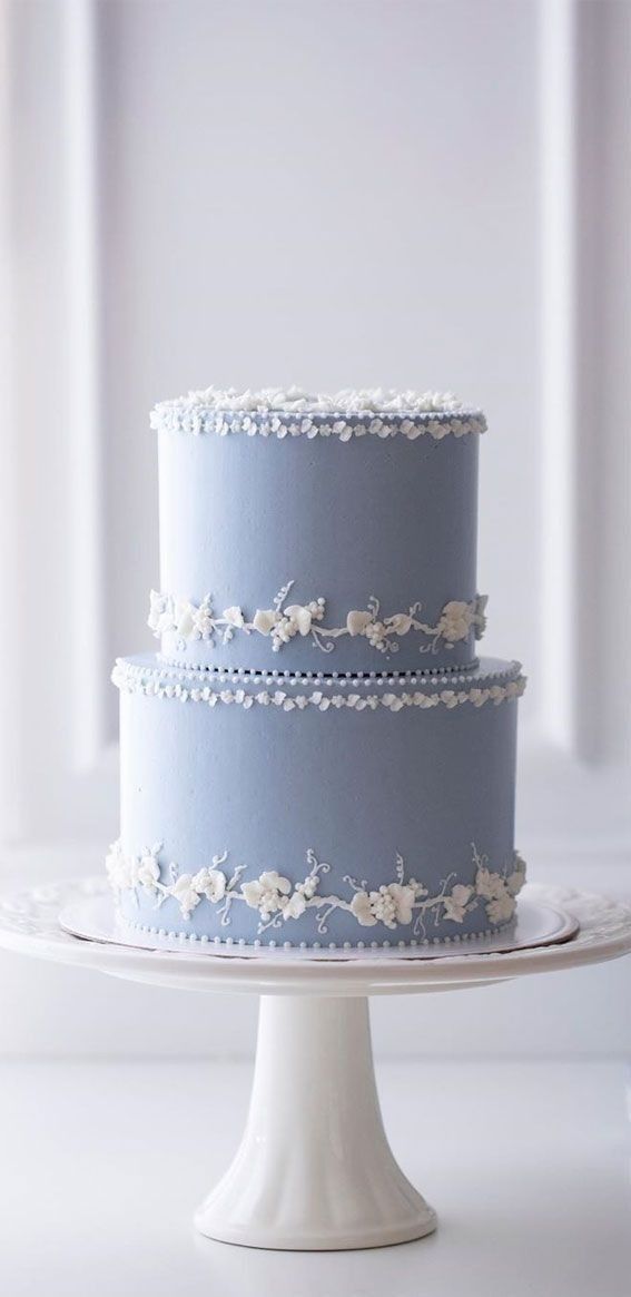 a three tiered blue cake with white flowers on the top and bottom, sitting on a pedestal