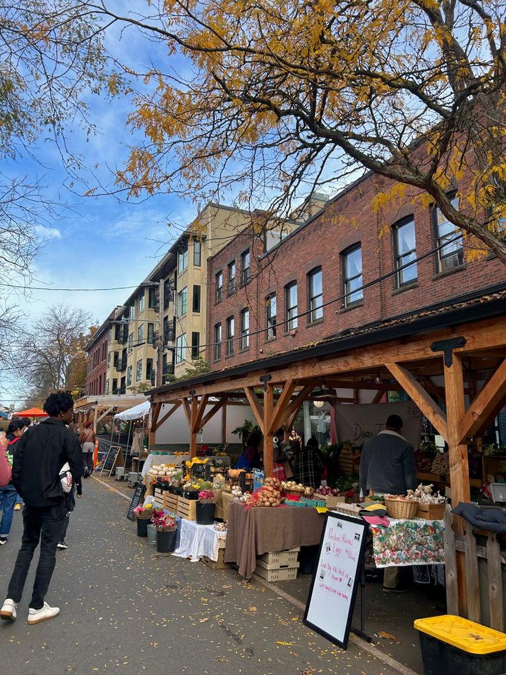 an outdoor farmers market with people shopping and selling items on the sidewalk near tall brick buildings