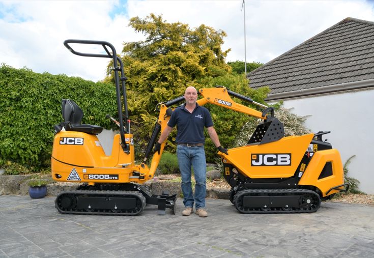 a man standing next to two small construction machines in front of a house with trees