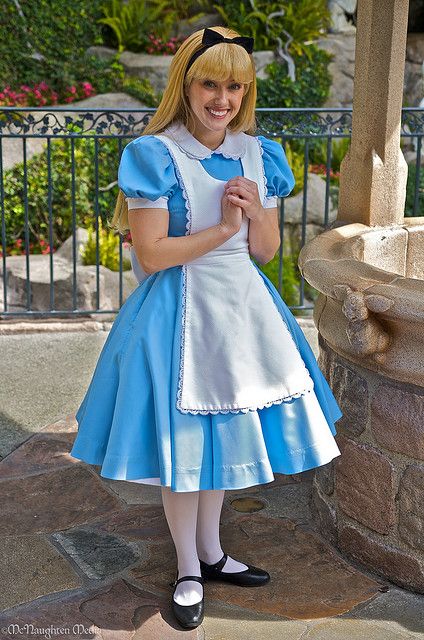 a woman dressed in a blue and white dress standing next to a fountain with her hand on her chest