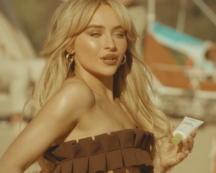 a woman in a brown dress is holding a small tube of sunscreen and looking at the camera