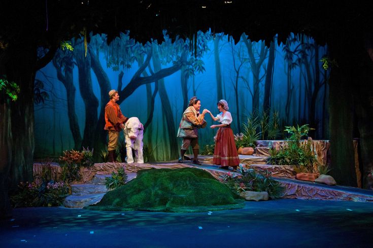 three people standing on stage in the middle of a forest with trees and rocks around them