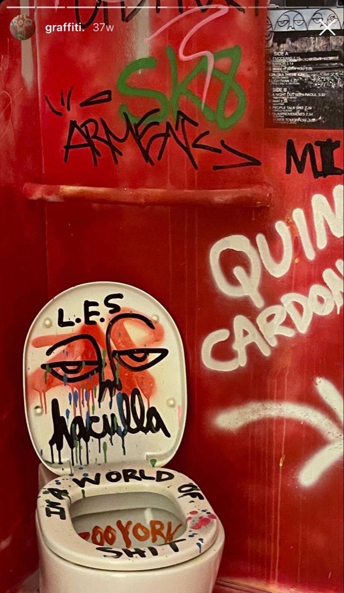 a toilet with graffiti on the lid in a bathroom stall, next to a red wall