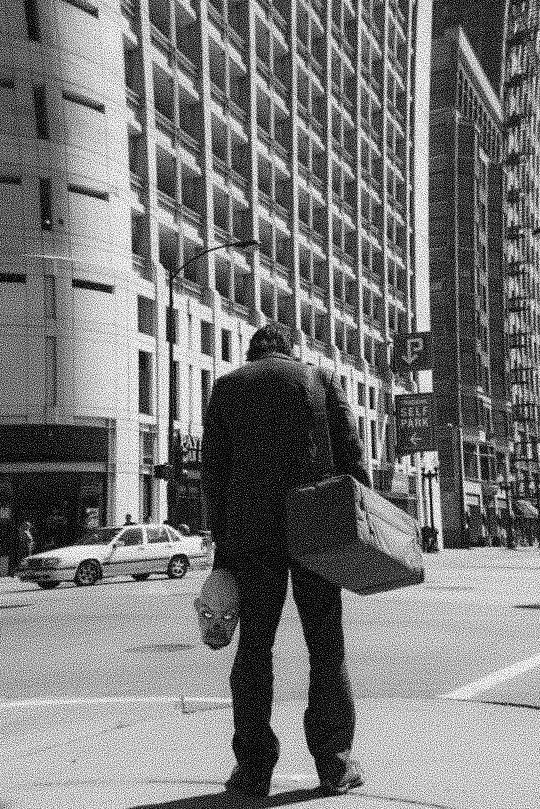 black and white photograph of man walking down the street with bags on his back, in front of tall buildings