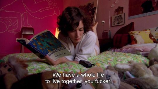 a woman laying in bed reading a book with stuffed animals on the floor next to her