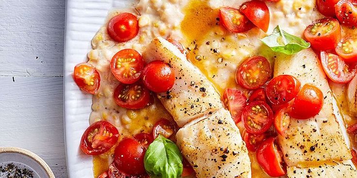 two fish fillets with tomatoes and basil on a white plate next to a bowl of seasoning