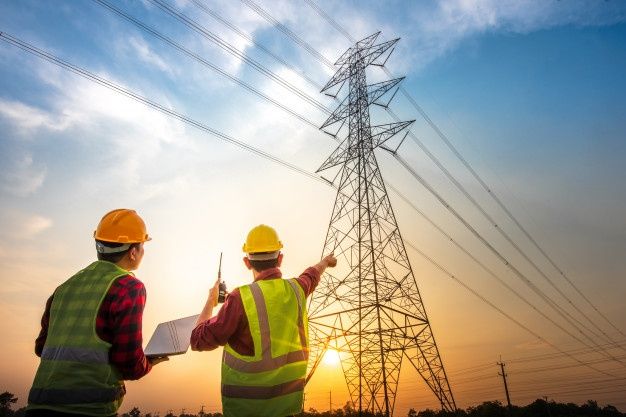 two men in safety vests and hard hats standing next to an electric pole with the sun setting behind them