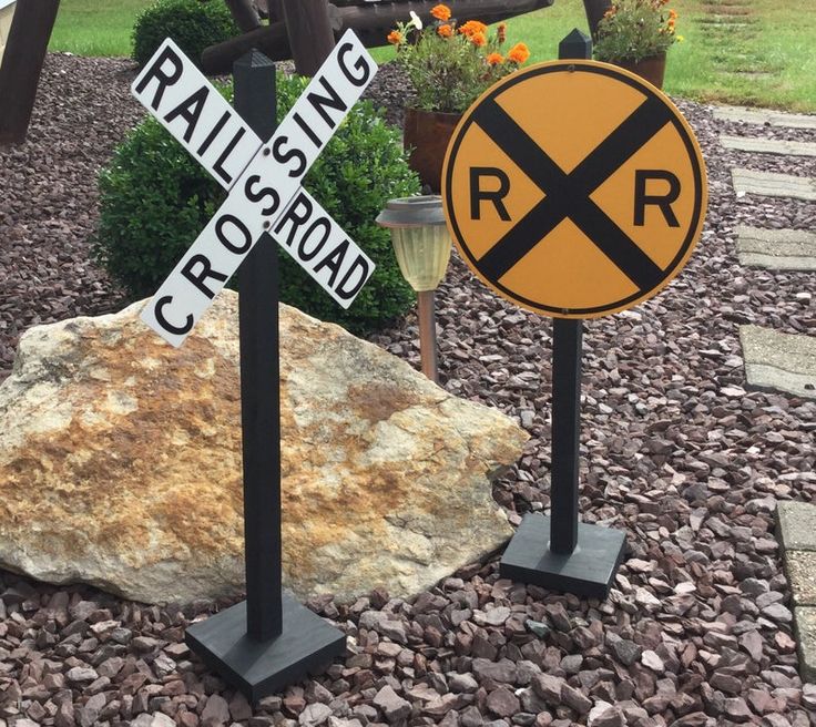 two railroad crossing signs sitting on top of rocks in front of a rock and flower garden