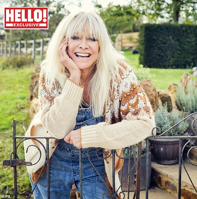 a woman with blonde hair and blue jeans leaning on a gate smiling at the camera