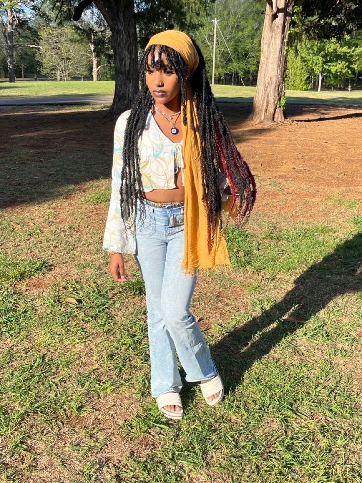 a woman with dreadlocks standing in the grass wearing jeans and a yellow scarf