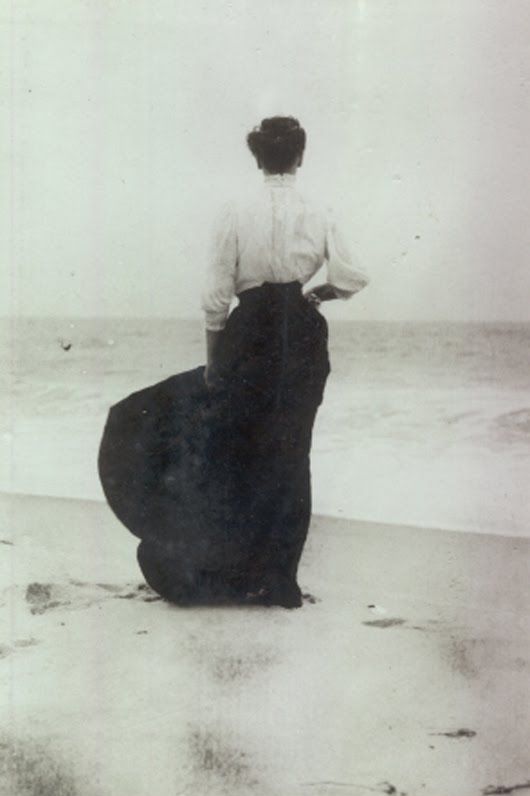 an old photo of a woman standing on the beach looking out at the ocean with her back to the camera