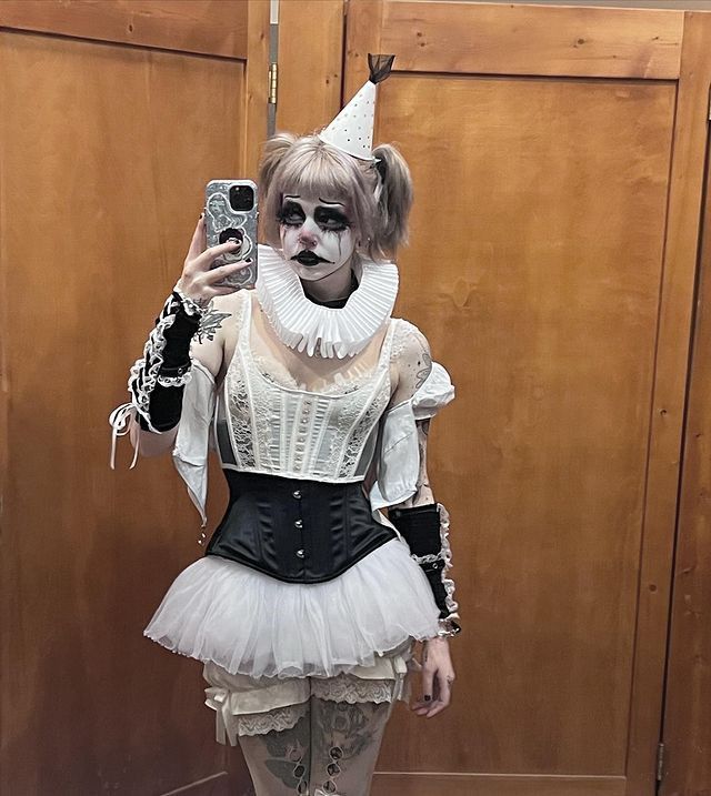 a woman dressed as a clown taking a selfie with her cell phone while standing in front of a wooden door