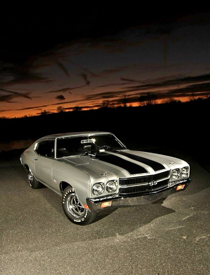 an old muscle car parked in a parking lot at night with the sun setting behind it