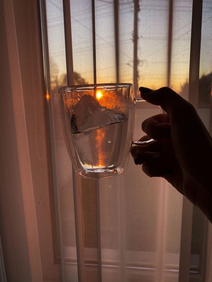 a hand holding a wine glass in front of a window with the sun going down