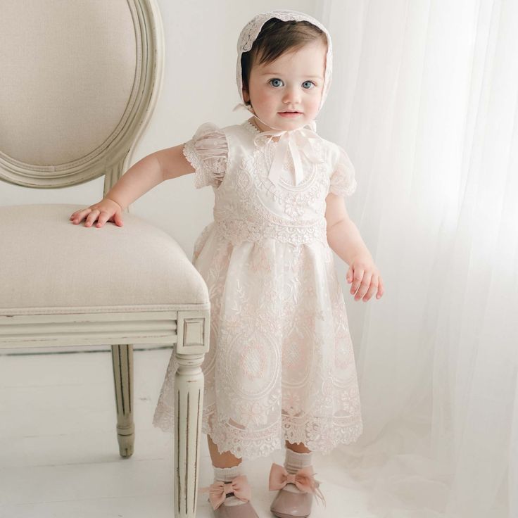 A toddler wearing the delicate, handmade Elizabeth Christening Dress with embroidered lace and a matching bonnet stands beside a vintage-style upholstered chair. The child has one hand resting on the chair and is looking slightly to the side, with soft natural light filtering through a sheer curtain in the background. Family Dress, Silk Dupioni, Baptism Dress, Dupioni Silk, Pink Champagne, Swiss Dot, Ivory Color, Embroidered Lace, Vintage Pink