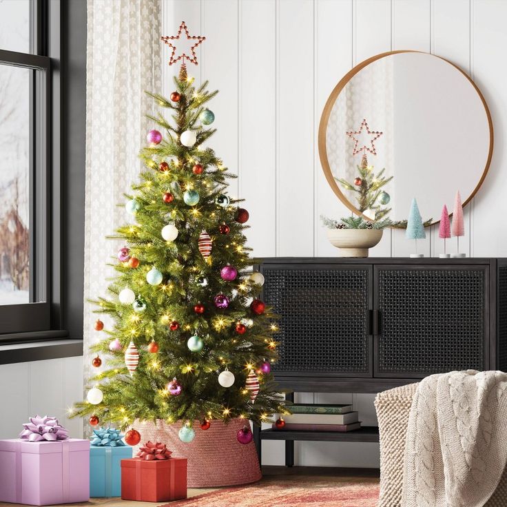 a decorated christmas tree in front of a window with presents under it and a round mirror on the wall