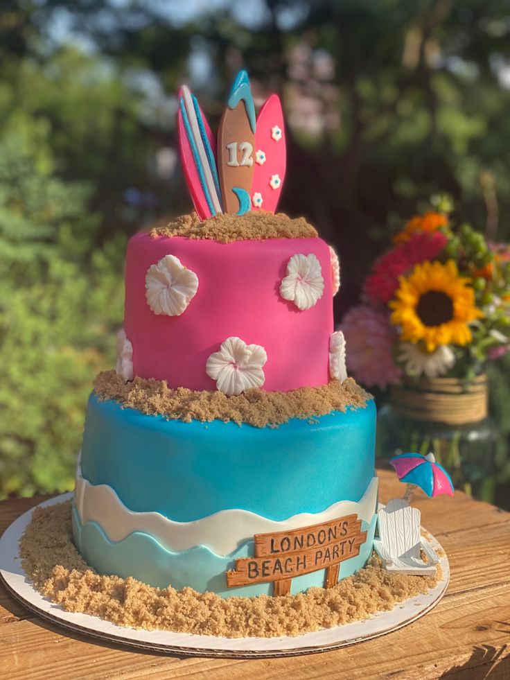 there is a pink and blue cake that has surfboards on it, with flowers in the background