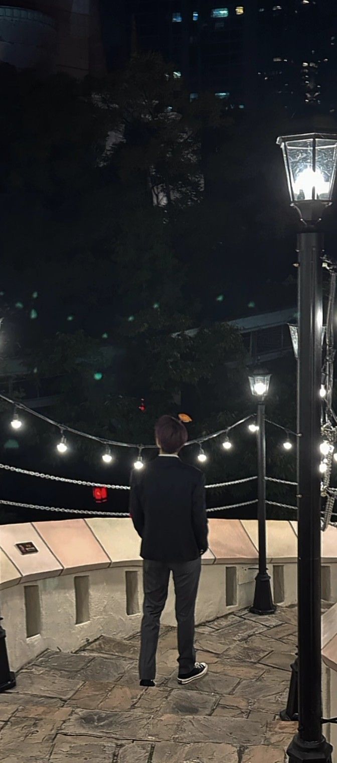 a man standing next to a lamp post on a sidewalk at night with lights strung across the street