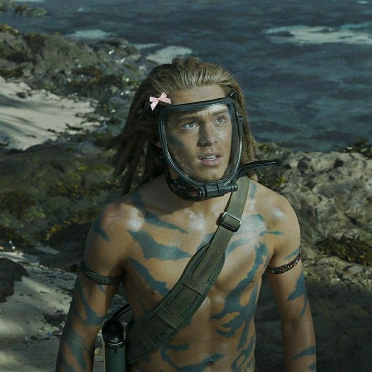 a man with long hair wearing a helmet and camouflage paint on his body is standing near the ocean