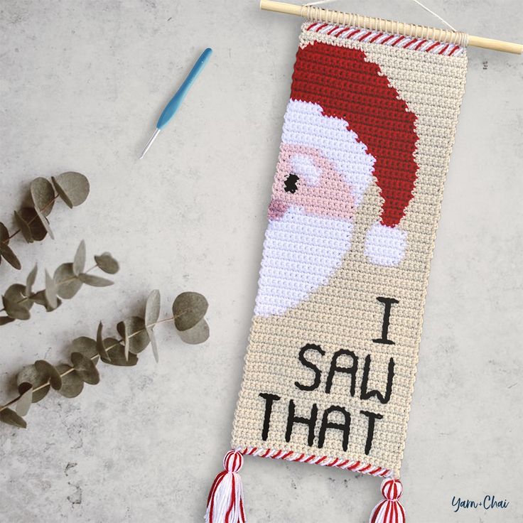 a cross stitch santa clause hanging on a wall next to some plants and needles with the words i say that