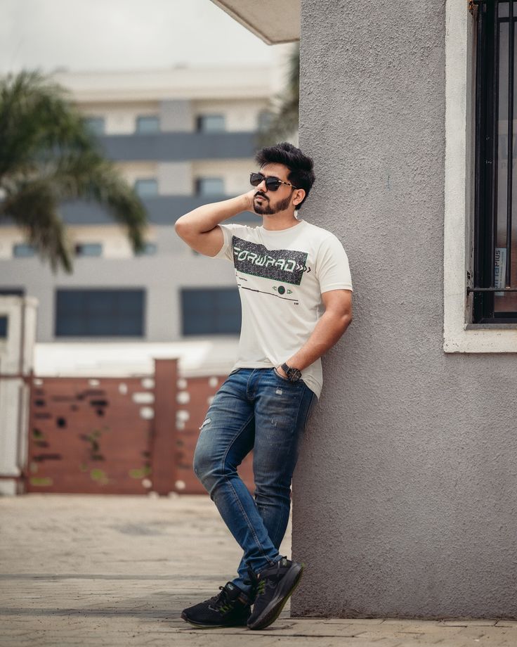 a man leaning against a wall with his hand on his head and wearing sunglasses, standing in front of a building