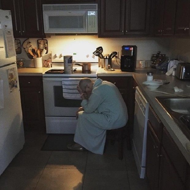 an old man sitting in a kitchen with his head on the stove and looking at something