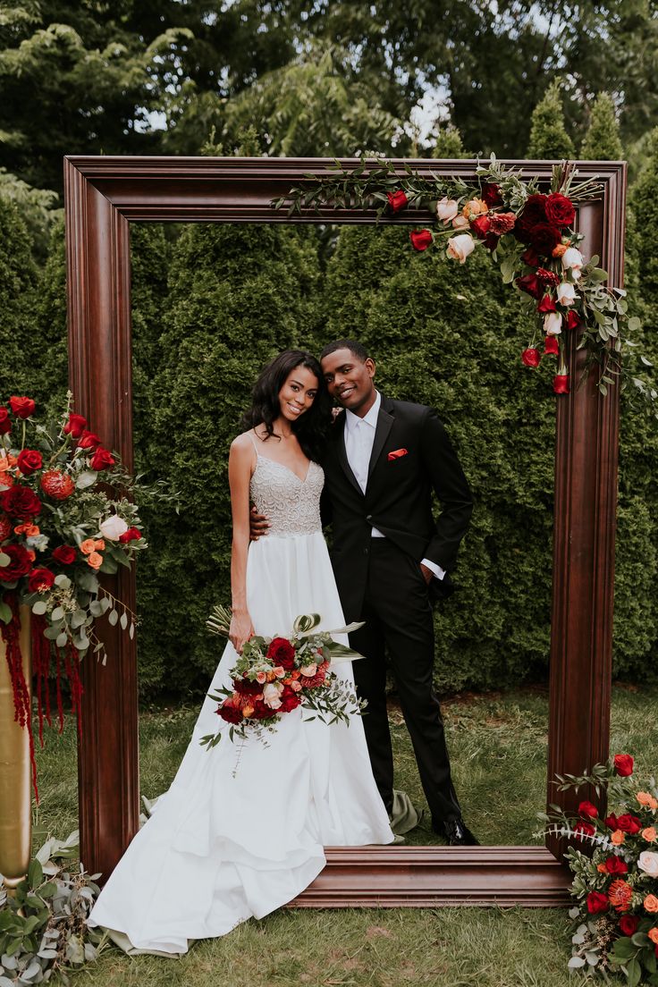 a bride and groom standing in front of a wooden frame with red flowers on it