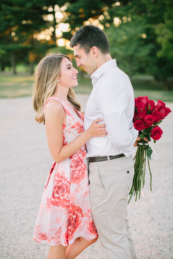 a man and woman standing next to each other holding red roses in their hands while the sun is setting behind them