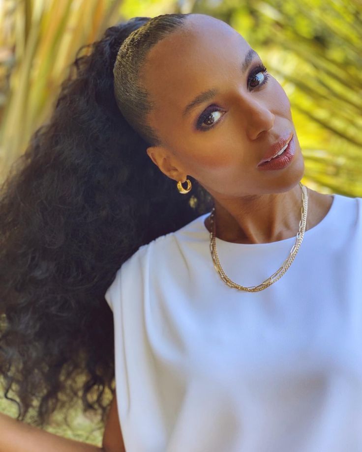 a woman with long black hair wearing a white shirt and gold necklace, looking at the camera