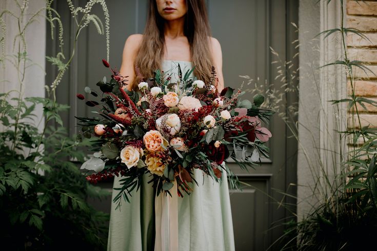 a woman standing in front of a door holding a bouquet of flowers and greenery