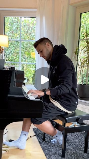 a man sitting at a black piano in front of a window and playing the piano