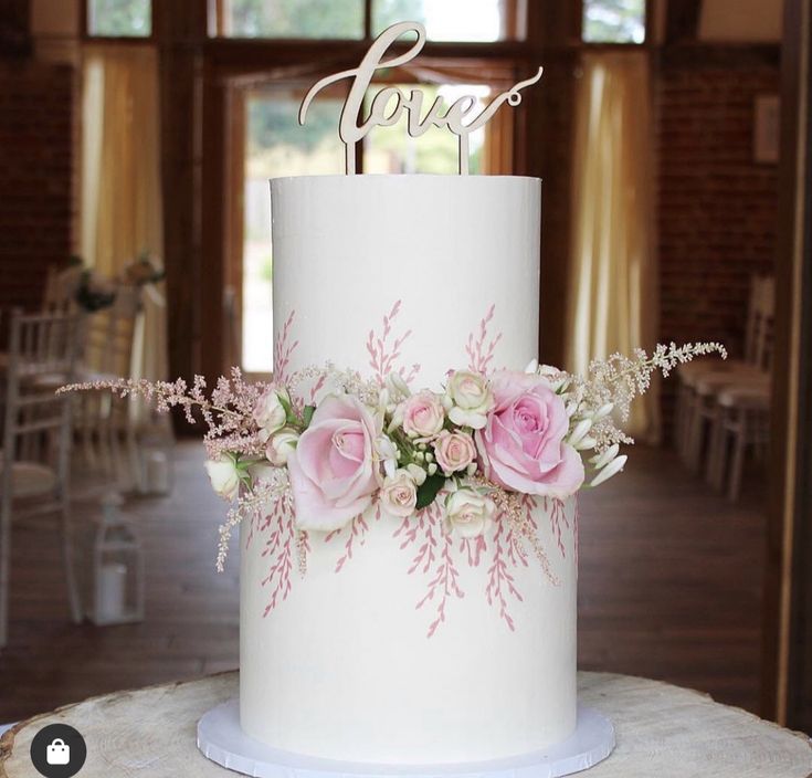 a white wedding cake with pink flowers and the word love on top is sitting on a tree stump