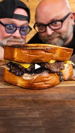 two men sitting next to each other in front of a sandwich on a cutting board